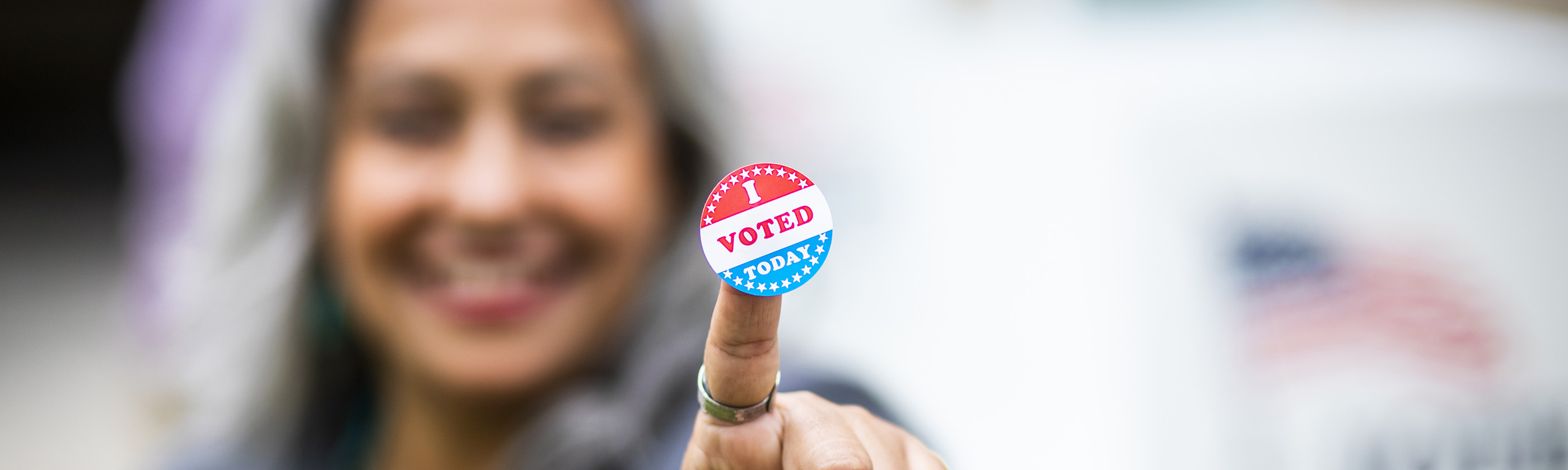 A woman in soft focus holds a sticker toward the camera. The sticker reads I voted today.