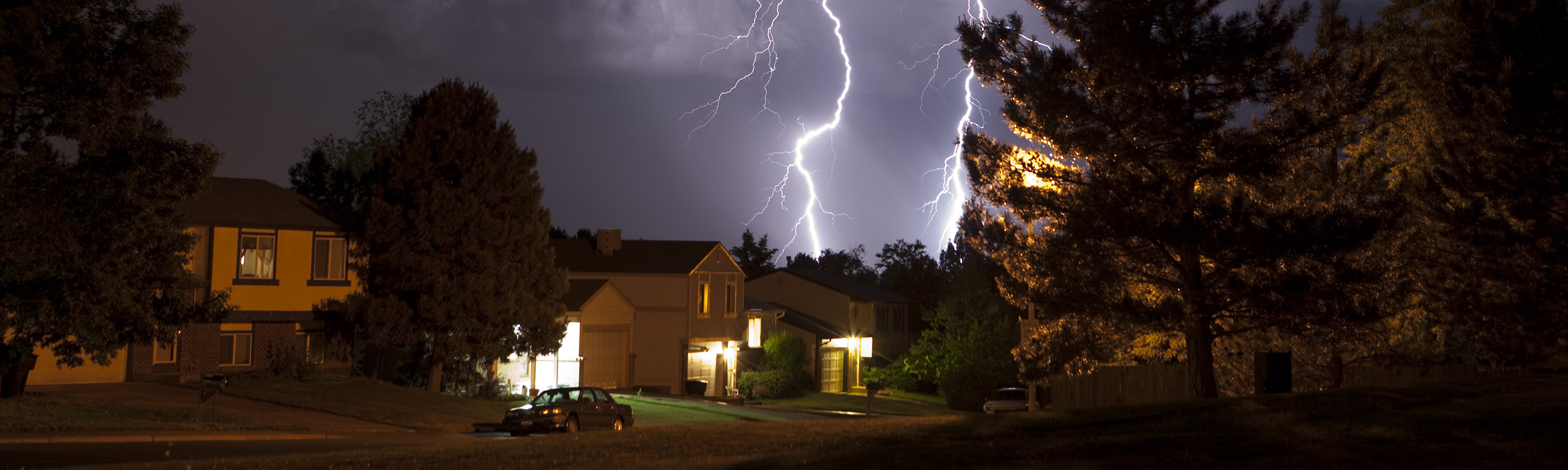 Lightning from a thunderstorm strikes behind a row of houses in a suburban neighborhood.